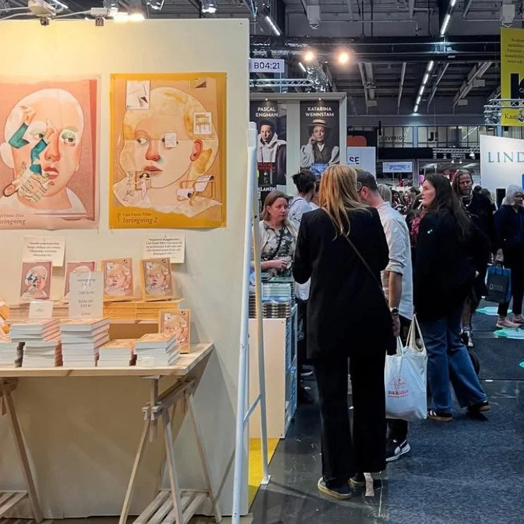 En bokmässa med en monter som har konstverk av en flicka och en pojke på väggen och flera böcker staplade på ett bord. Människor som går och tittar på montrarna.