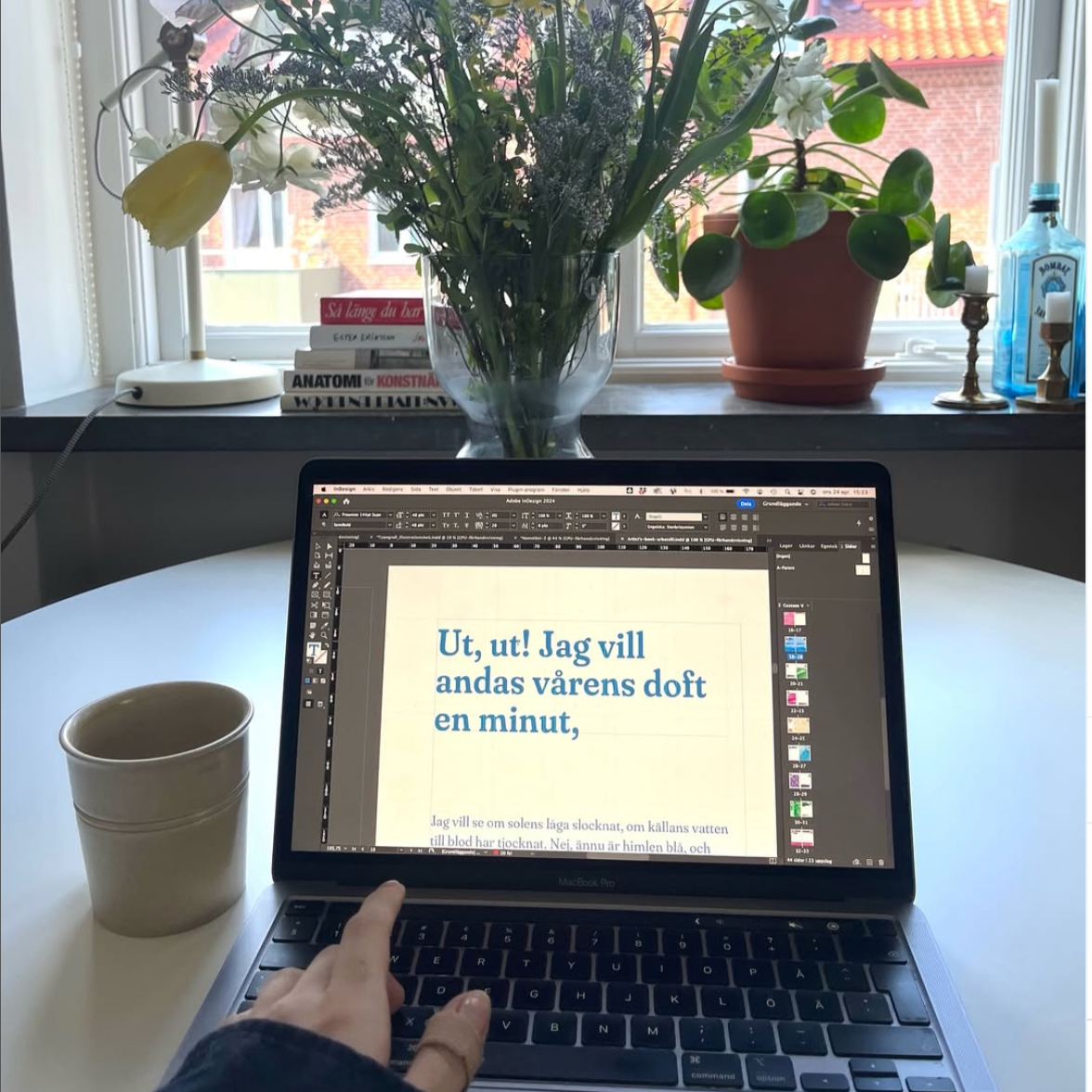 En laptop på ett runt bord med en hand som pekar på skärmen, en tom mugg bredvid, i bakgrunden en fönsterbräda med blommor, böcker och två ljusstakar.