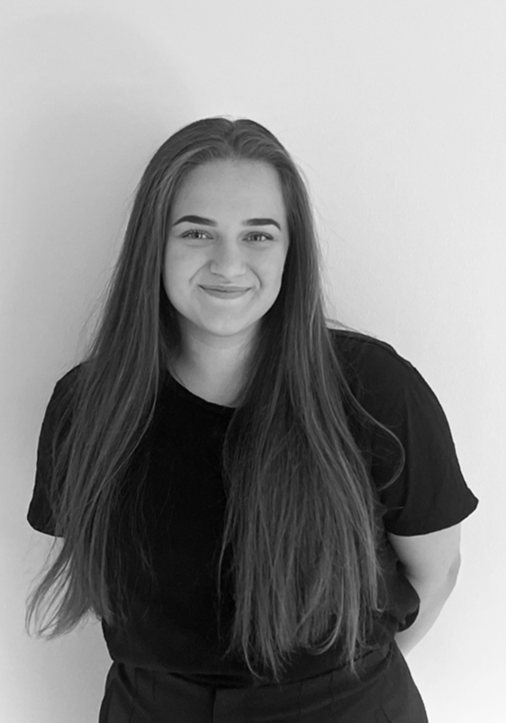 Ung kvinna med långt hår som ler, bär en svart T-shirt och står mot en ljus vägg.