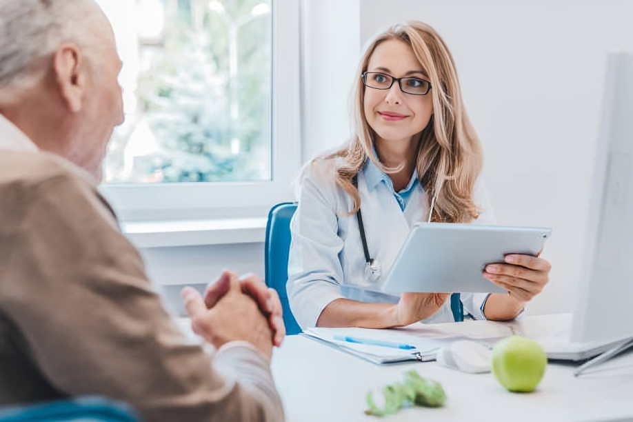 A young female doctor in a white coat and black glasses sitting at her desk, holding a tablet, smiling, and talking to an older male patient in a beige coat, in a bright medical office with a large window, an apple, and a toy dinosaur on the desk.