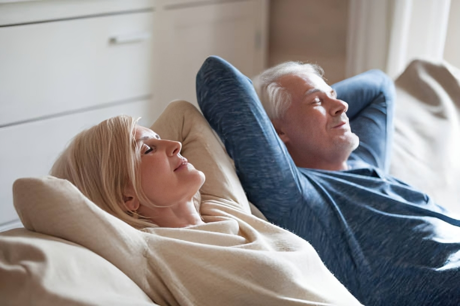 An older man and woman relaxing on a sofa with eyes closed, leaning on pillows, enjoying peaceful moment.