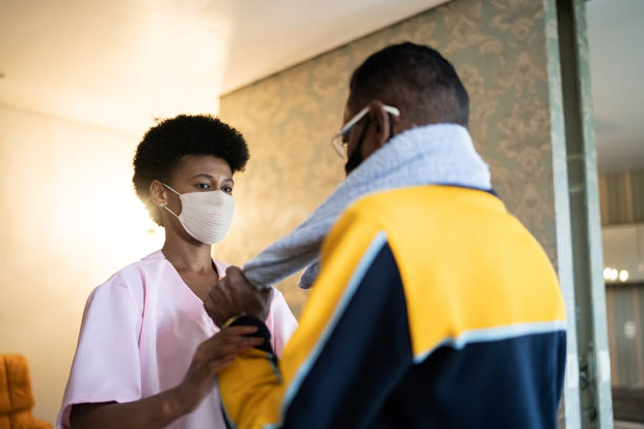 A woman in pink scrubs helping a man adjust his face mask, both wearing masks, indoors with warm lighting.