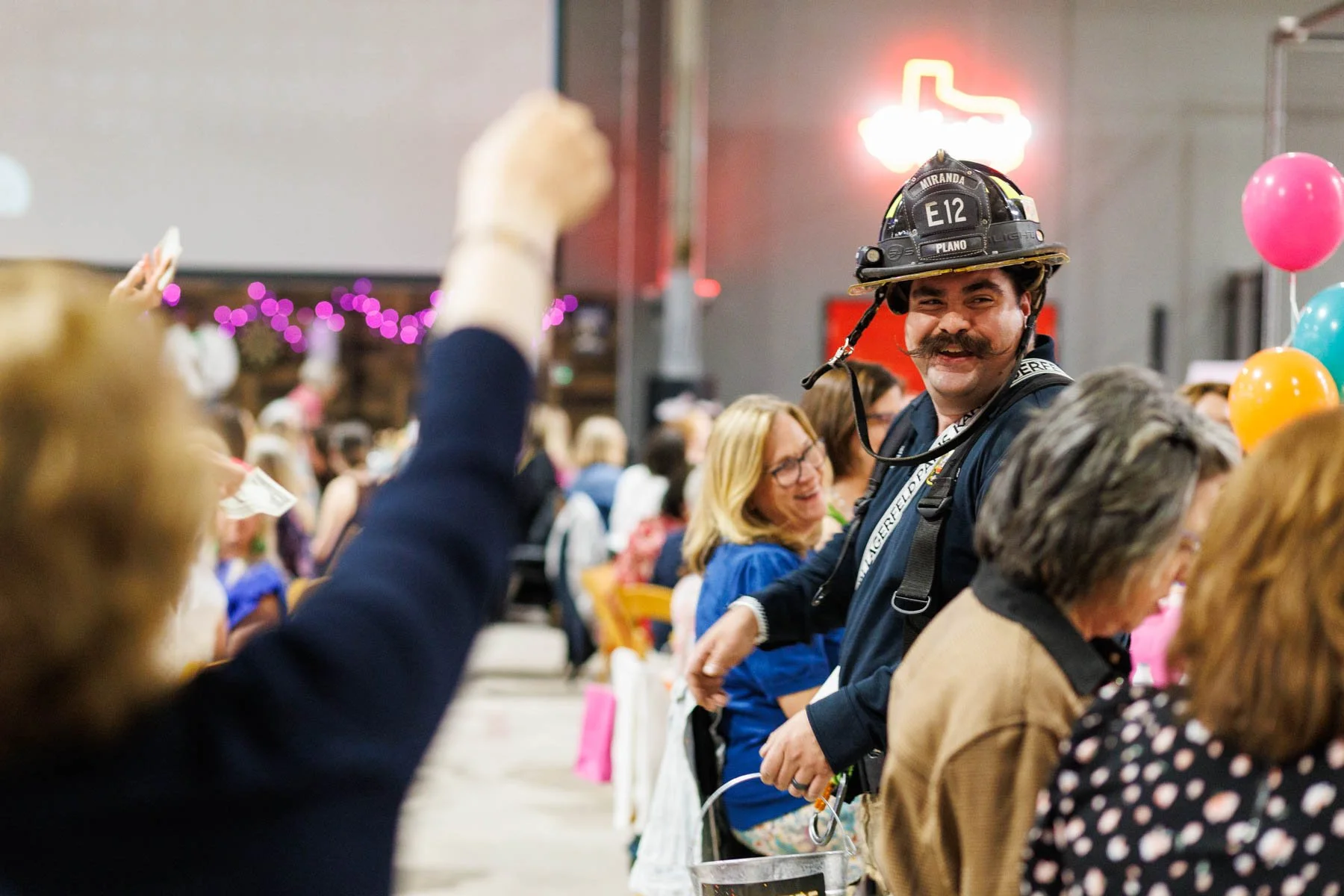A firefighter with a mustache, wearing a black helmet and navy uniform, stands smiling at a party or celebration with balloons and people in the background.