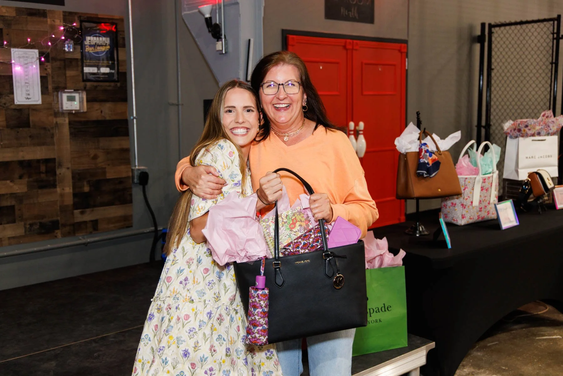 Two smiling women hugging at a gift or craft show, one woman holding a black purse with gift tissue paper inside, and bags of purses and gifts displayed on a table behind them.