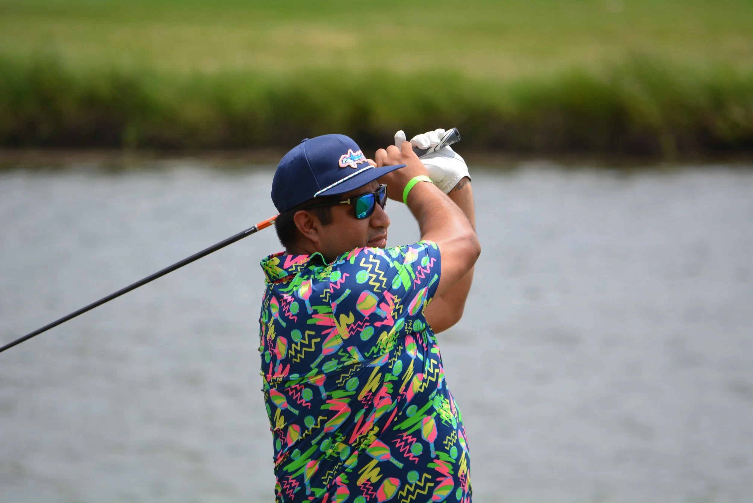 Man in colorful shirt and blue cap playing golf by a body of water, holding a golf club.