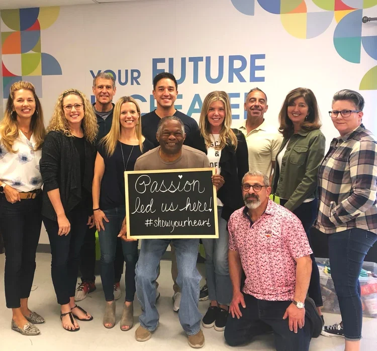 Group of eleven people standing in front of a wall with a motivational message, holding a sign that says "Passion led us here! #showyourheart".