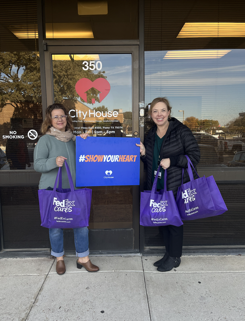 Two women standing outside a building holding purpleFedEx bags and a blue sign that reads #SHOWYOURHEART. The building has a window with a heart-shaped logo and the words 'City House' at 350, with a notice for office hours.