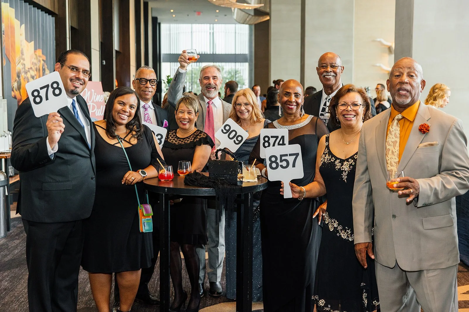 Group of people at a social event holding auction paddles and drinks, dressed in formal attire, in a modern venue with large windows.