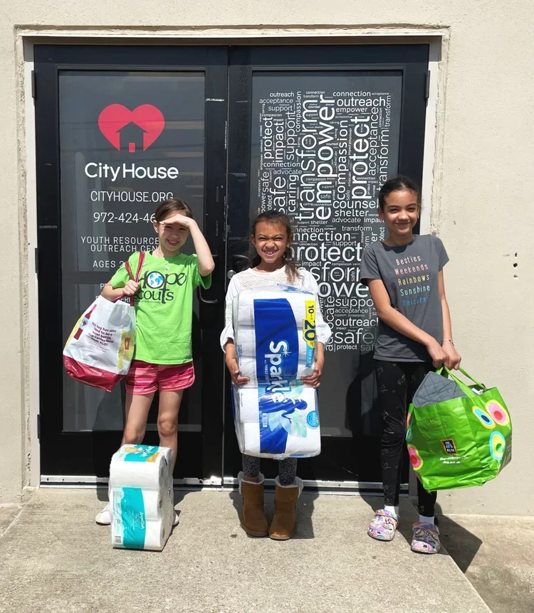 Three young girls standing outside a building with a door that has a Heart and House logo and the words "City House" and website info. They are smiling and holding grocery and paper towel bags. One girl is shielding her eyes from the sun. The door's glass also has a word cloud.