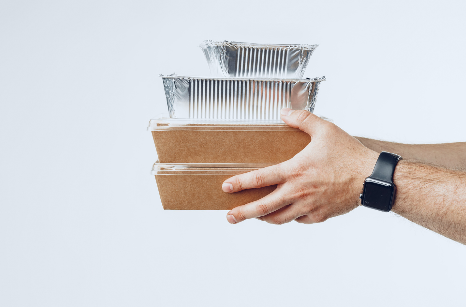 A person holding three stacked takeout food containers, including two aluminum foil trays and a cardboard box, with a smartwatch on their wrist.