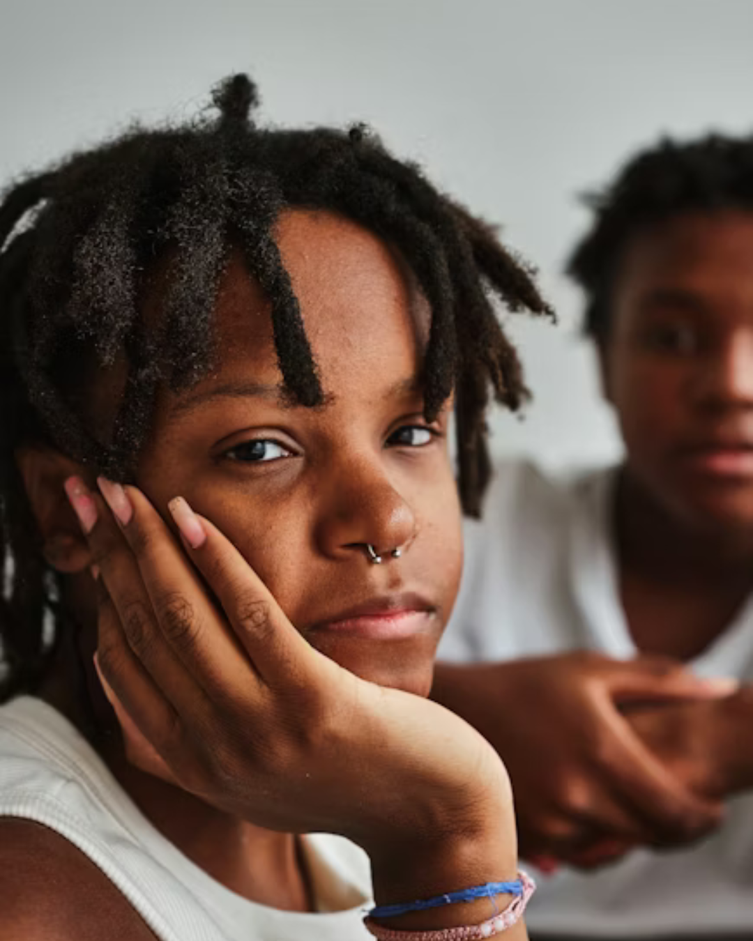 Close-up of a young woman with natural dreadlocks, wearing a septum piercing, resting her chin on her hand and looking at the camera. A young man with dreadlocks is blurred in the background, resting his chin on his hand.