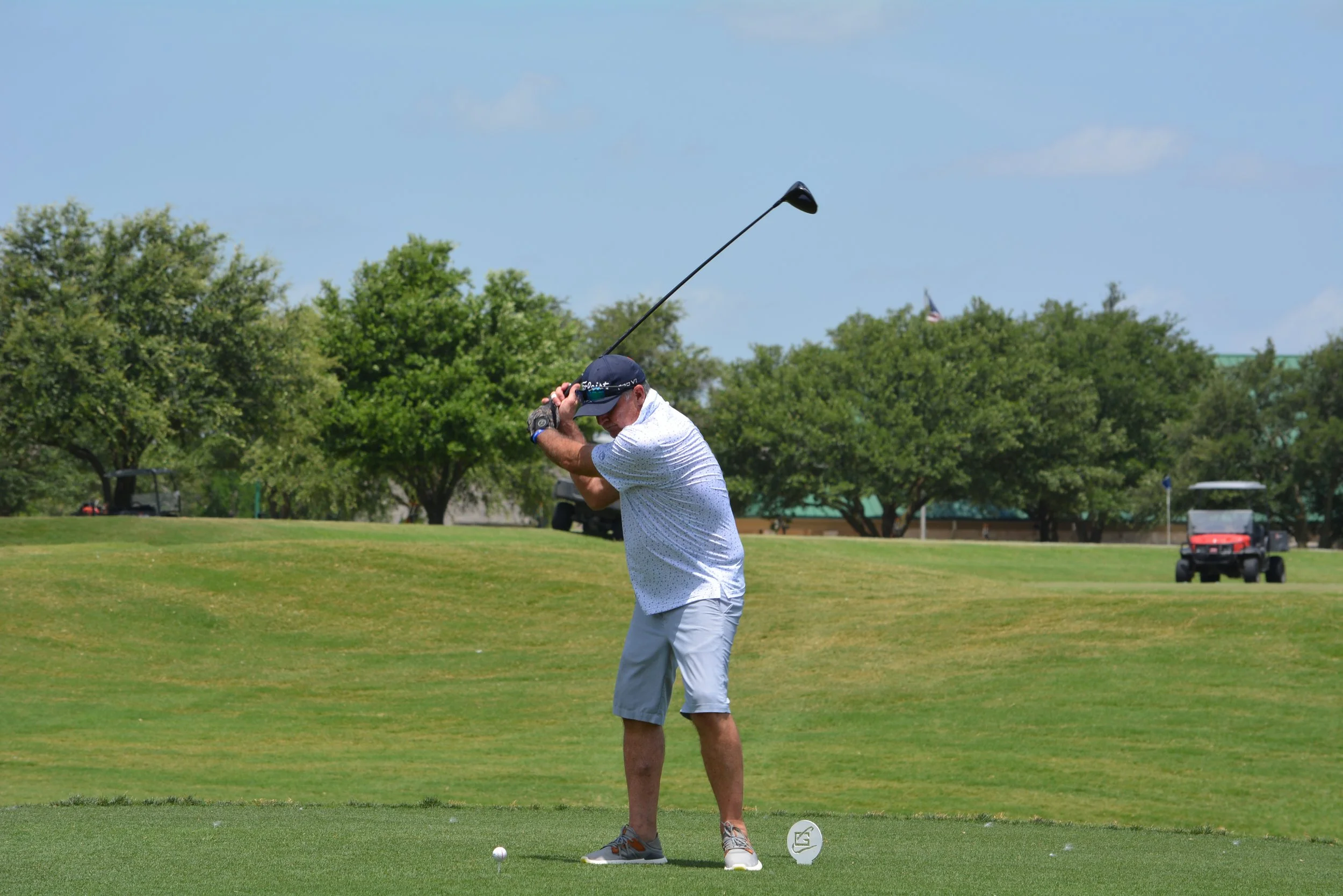 A man playing golf on a course, swinging a club, with trees and golf carts in the background.