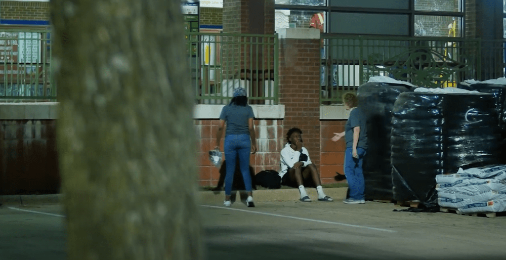 Three people talking to a person sitting on the sidewalk near stacks of black wrapped pallets outside a building.