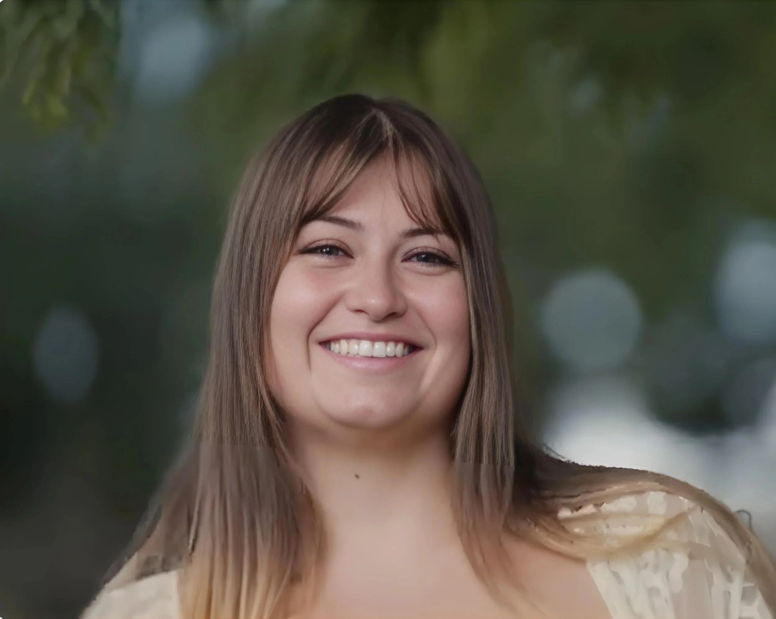 Close-up of a smiling young woman with long brown hair and a light-colored top, standing outdoors with a blurry green background.
