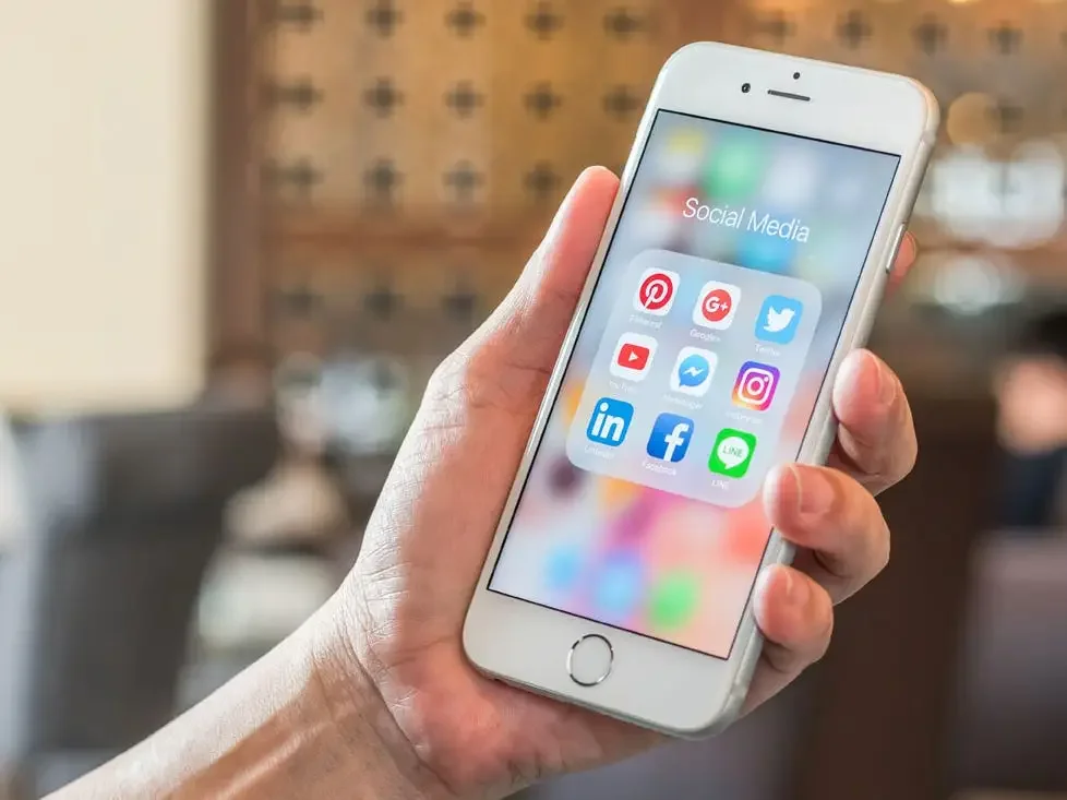 Hand holding a white smartphone displaying social media app icons in a folder labeled 'Social Media'.