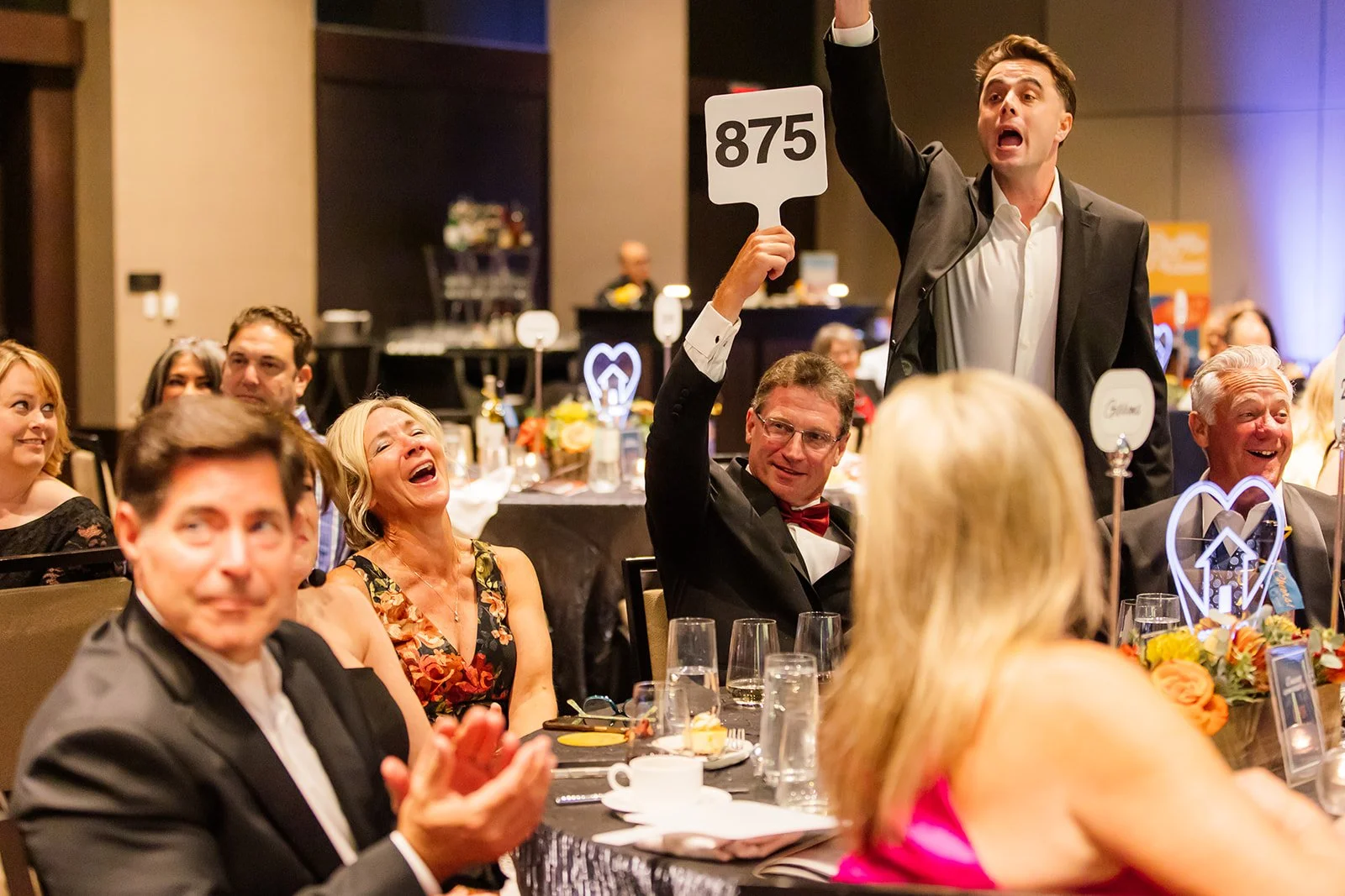 People dressed in formal attire at a banquet, a man standing and raising a paddle with the number 875, others are seated, some clapping and laughing, decorated tables with flowers and glasses.