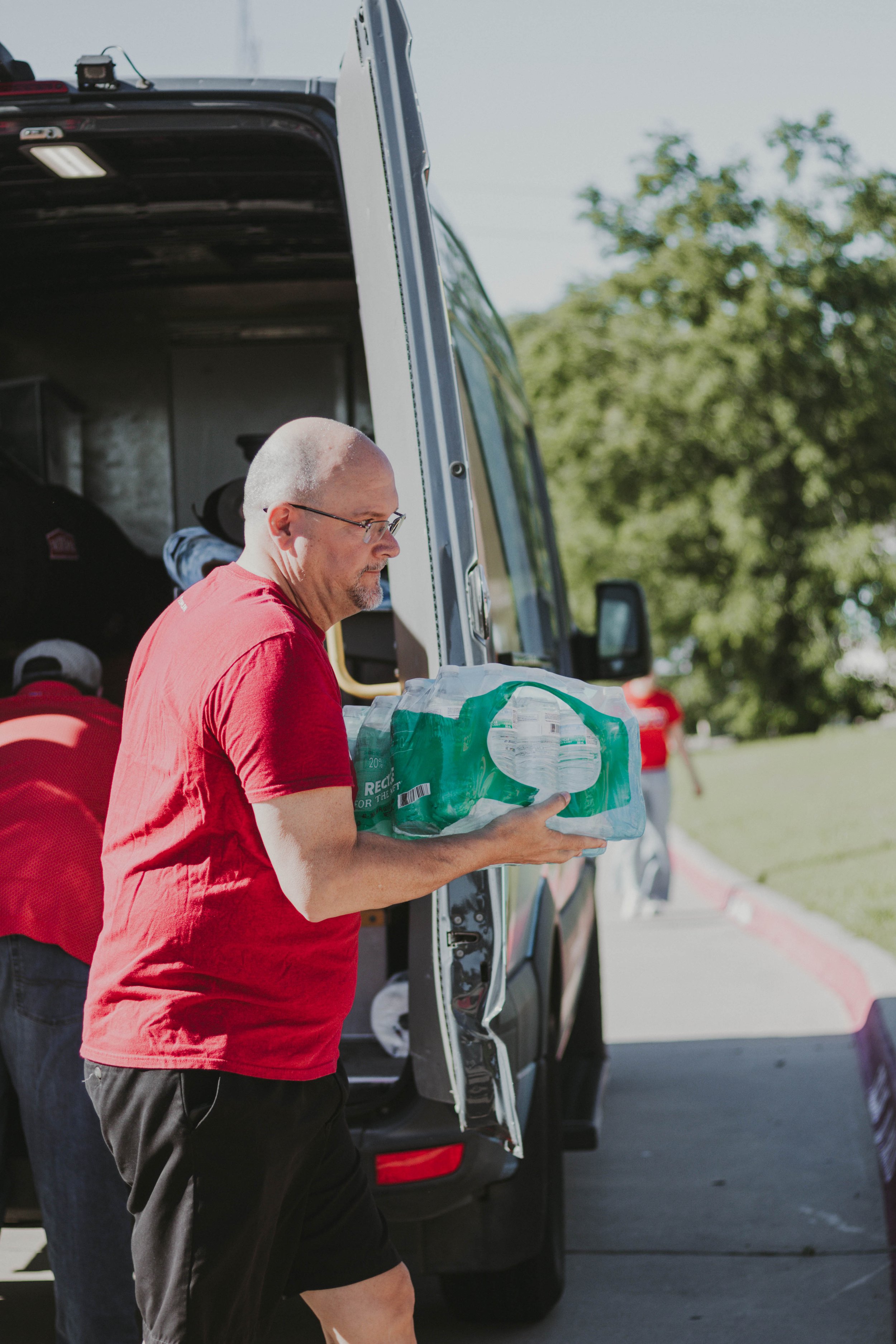 A man wearing a red shirt and black shorts is taking water bottles out of a van.