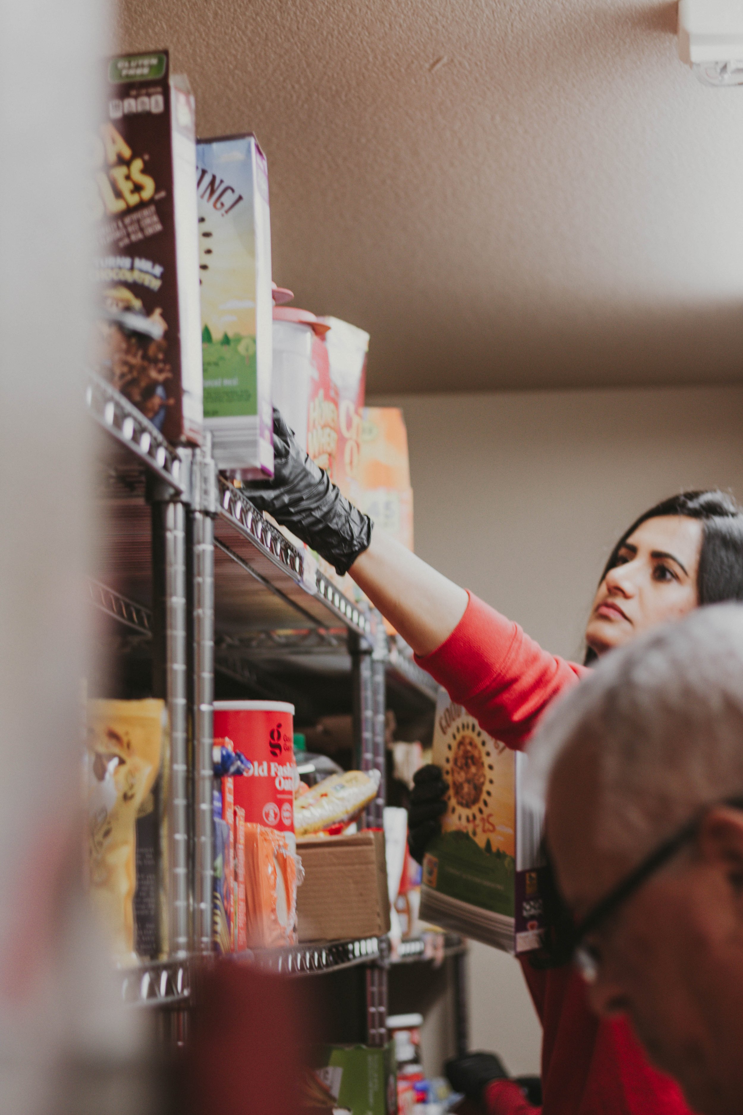A woman wearing a red shirt and black gloves reaching for food items on a metal shelf in a pantry or storage room.