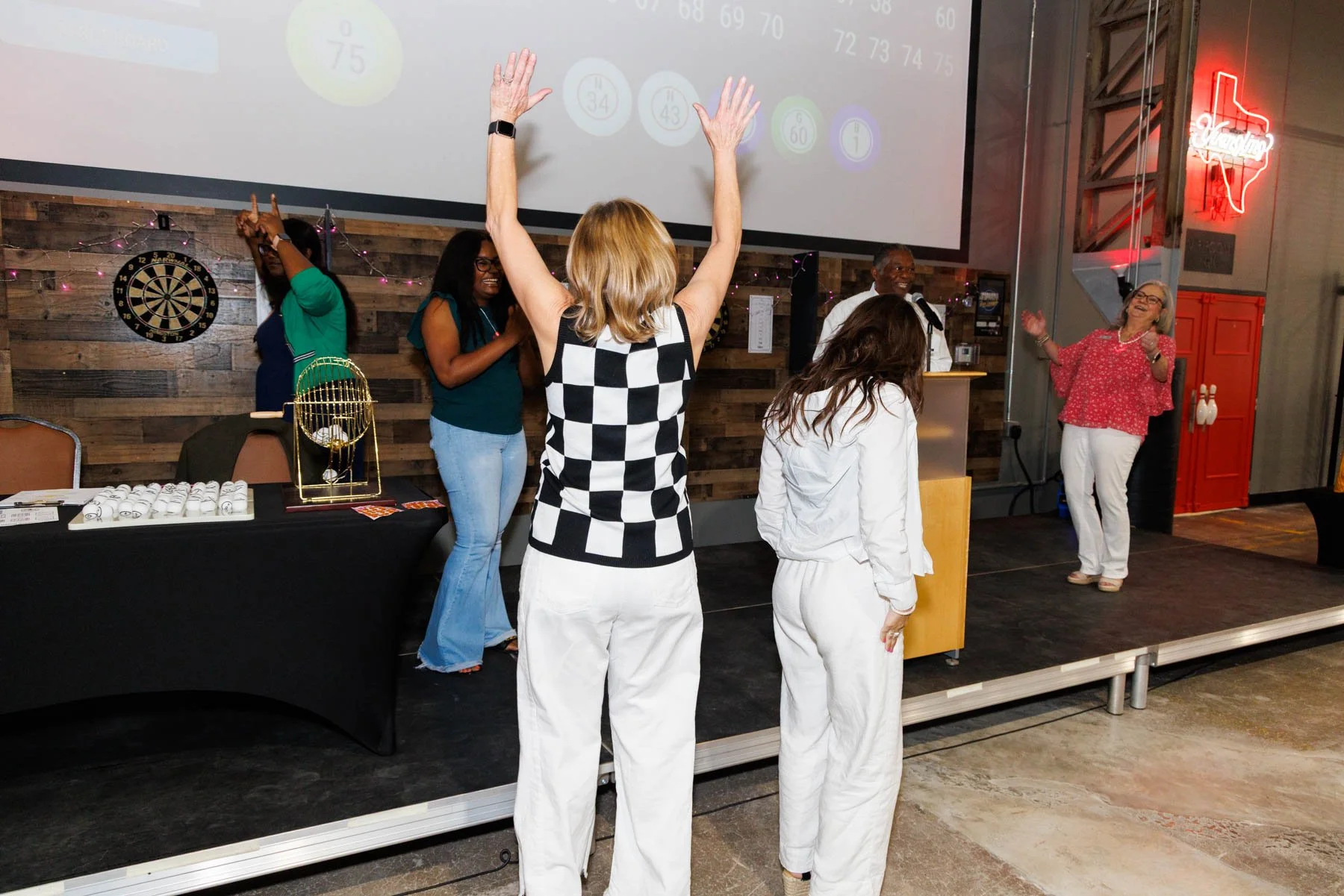 People dancing and enjoying music at a social event in a venue with wooden walls, dartboard, and neon Texas sign.