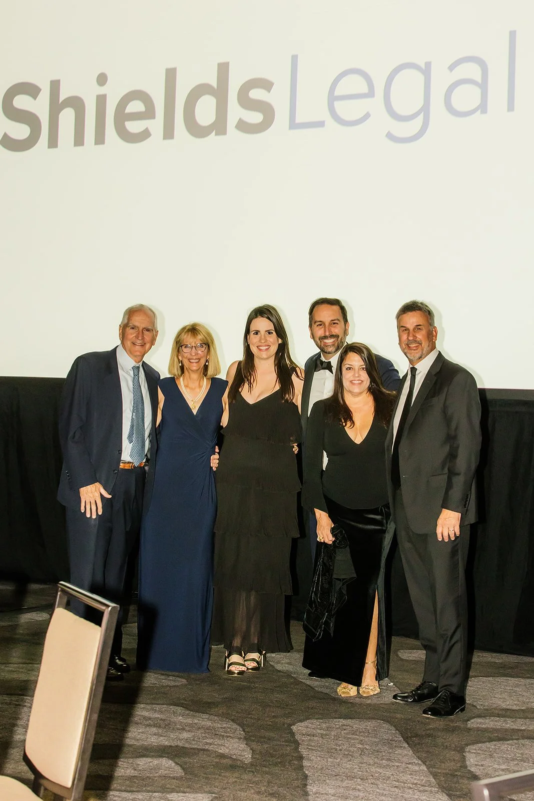 Group of six people dressed in formal attire standing together at an event with a large screen in the background displaying part of the word "Legal".