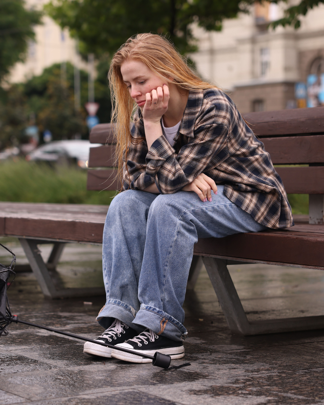A young woman with red hair sitting on a park bench with her chin resting on her hand, looking down with a worried expression. She is wearing a plaid shirt, jeans, and sneakers, and there is a launcher with a rubber band on the ground near her feet.