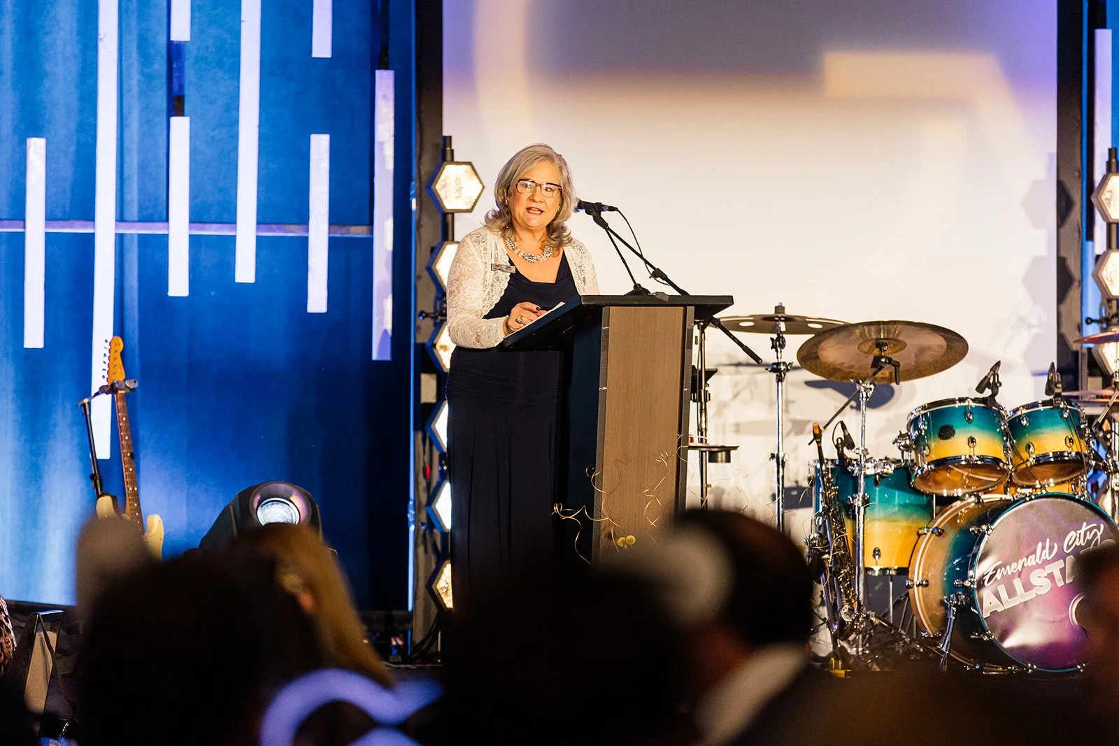 A woman with gray hair and glasses speaking at a podium during an event, with a stage featuring musical instruments including a drum set and an electric guitar in the background.