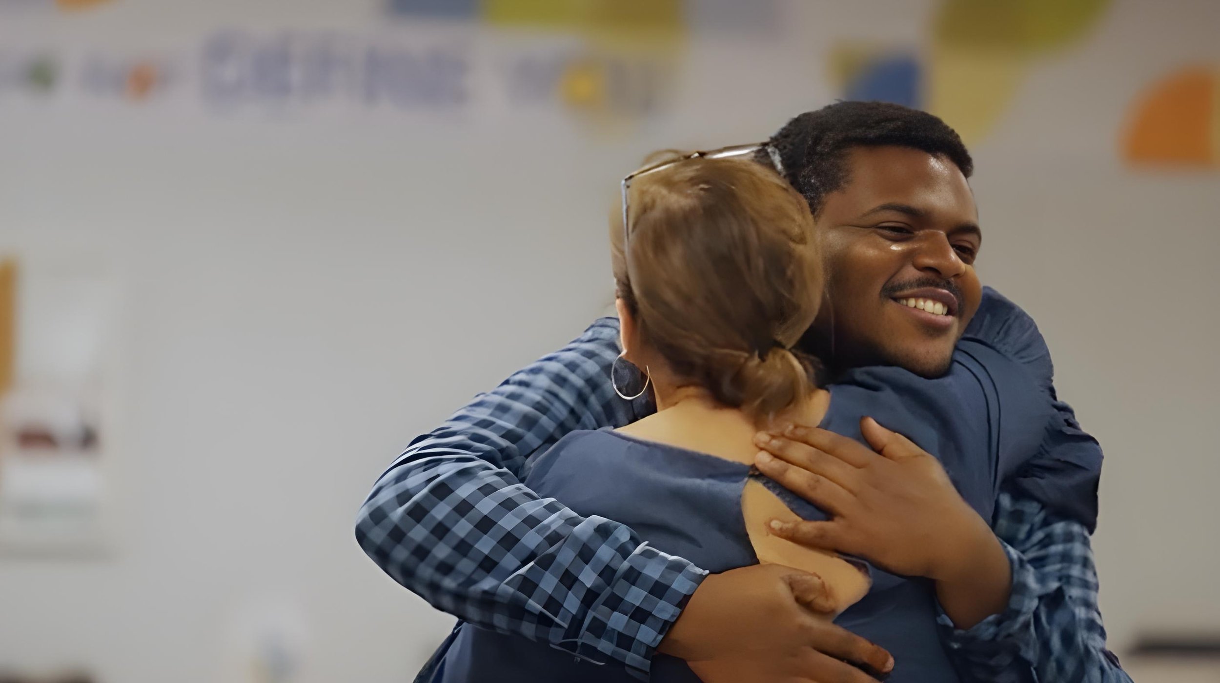A man and a woman hugging warmly and smiling in an indoor setting.