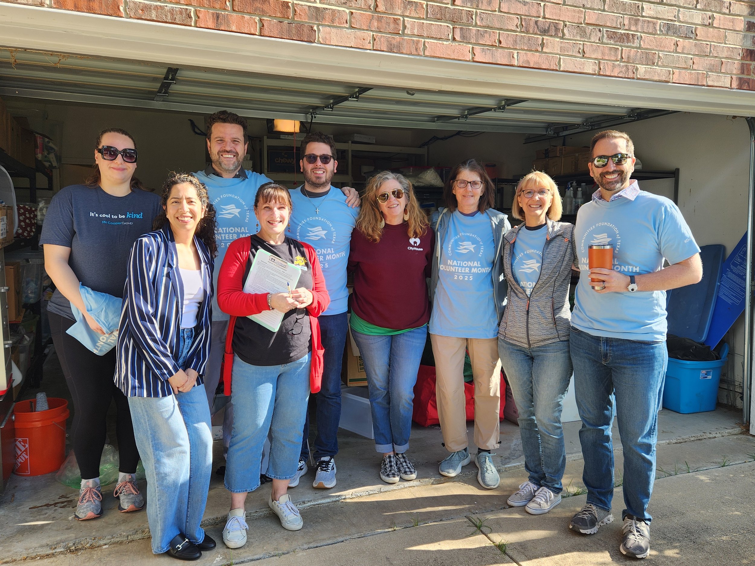 Group of nine people standing in front of a garage, smiling, some wearing matching blue event T-shirts that read 'National Volunteer Month 2025,' outdoors on a sunny day.