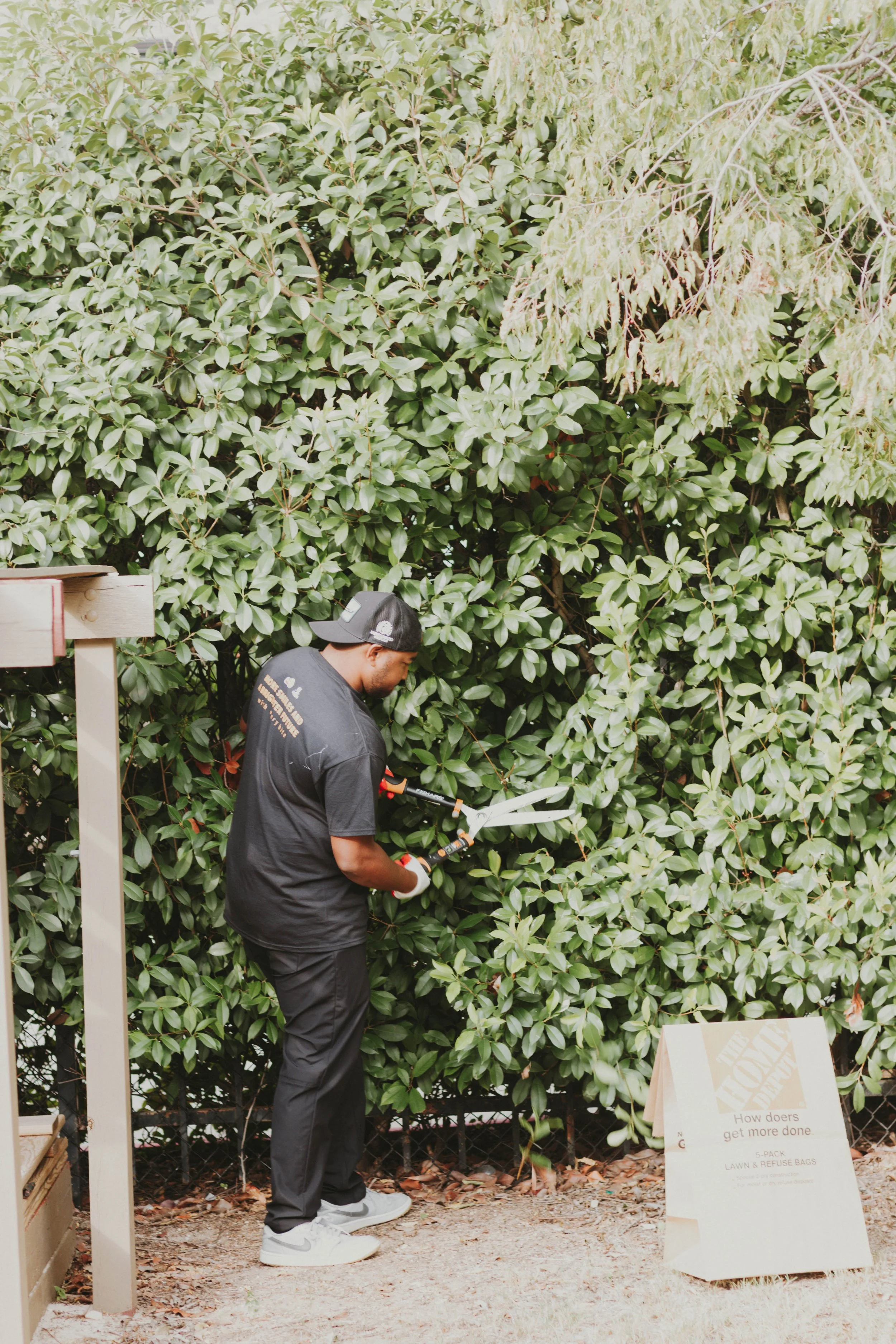 A man trimming bushes with hedge shears in a garden, wearing a black cap, black t-shirt, black pants, and gray shoes.