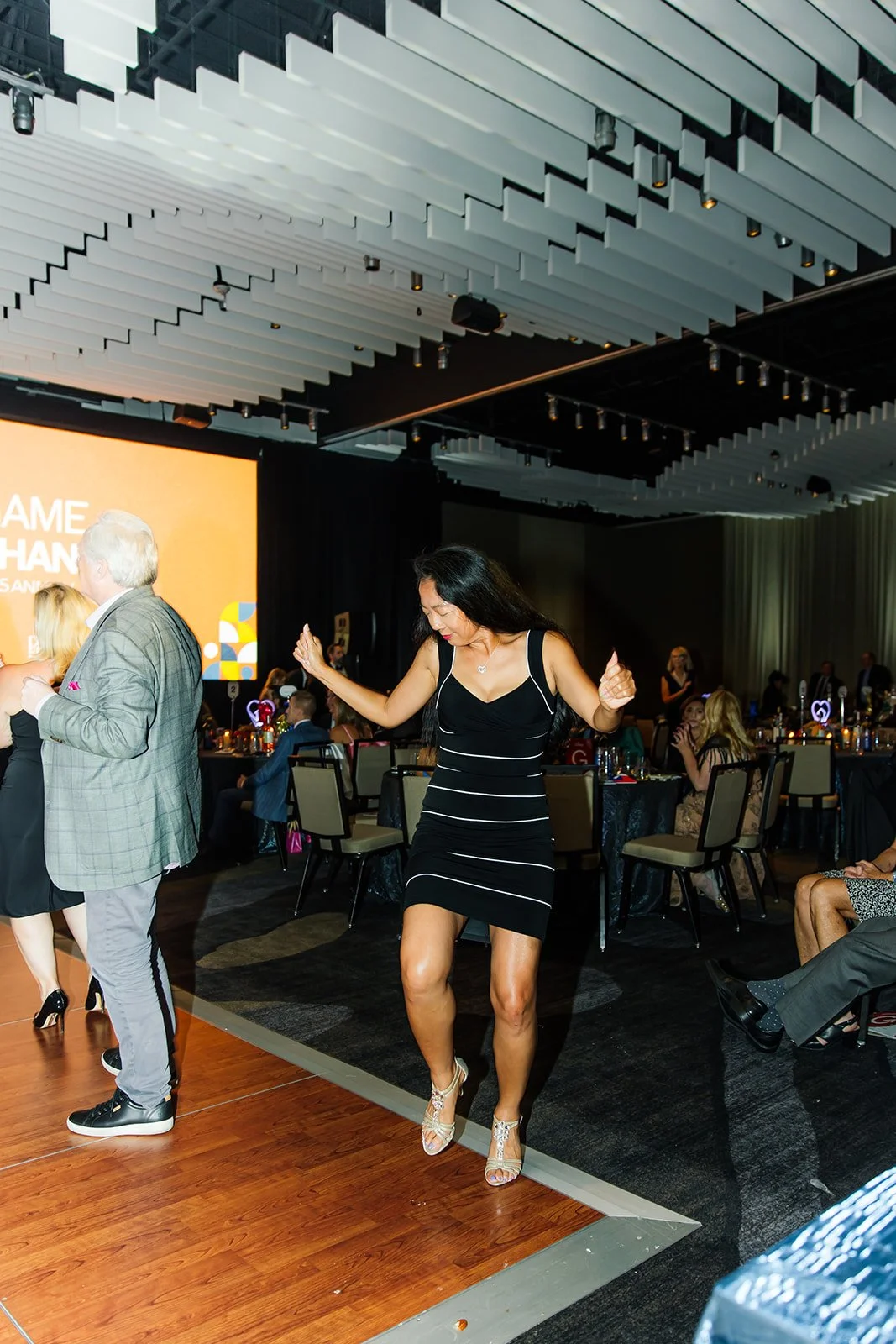 A woman in a black and white striped dress dancing on a wooden floor at an indoor event, with round tables and seated guests in the background.