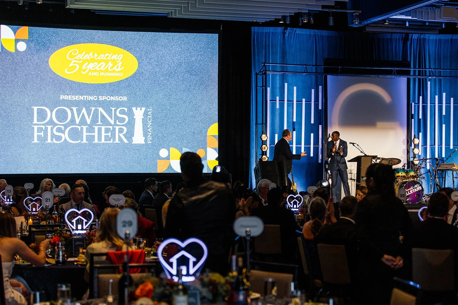 Event with a large screen displaying celebratory message and sponsor, a stage with two speakers, and audience members sitting at tables with illuminated heart-shaped signs.