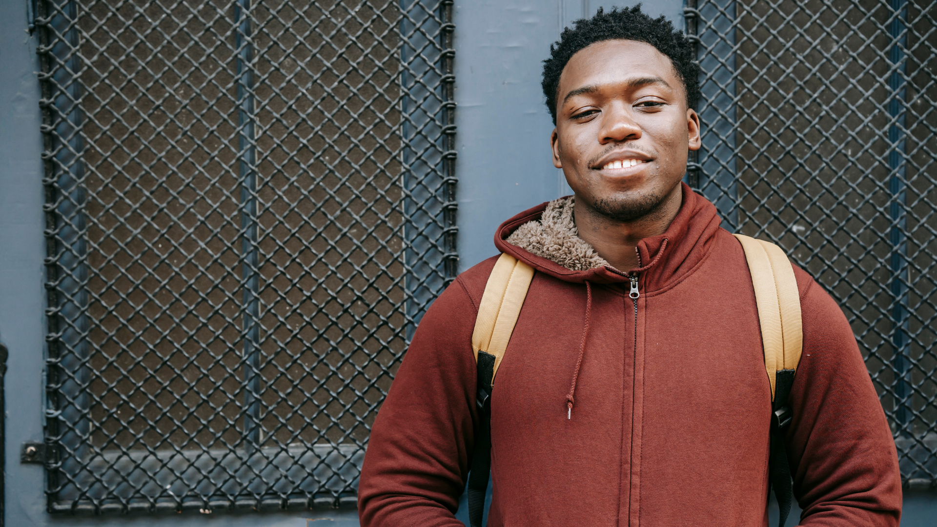 Young man with dark curly hair and a slight smile, wearing a brown hoodie with a furry lining and a yellow backpack, standing in front of a black metal fence.