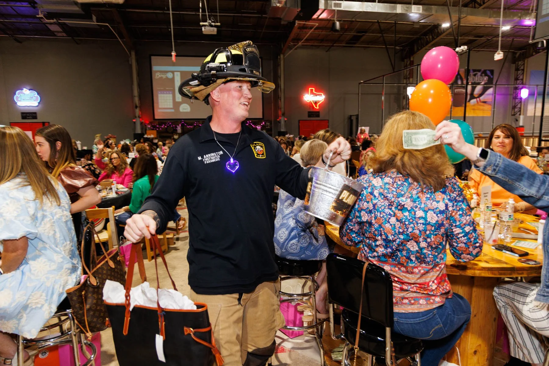 A firefighter wearing a helmet and a neon heart necklace is handing money to a woman with red hair, surrounded by people at a lively social event in a large indoor venue decorated with balloons.