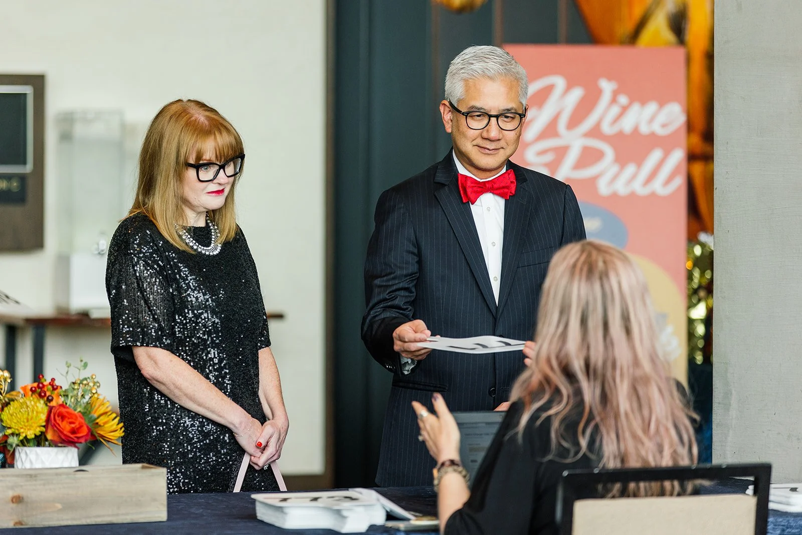 A man in a suit with a red bow tie is receiving a document from a woman sitting at a table, while a woman in black sequined dress and glasses observes. There is a sign in the background that reads 'Wine & Dull' and a flower arrangement on the table.