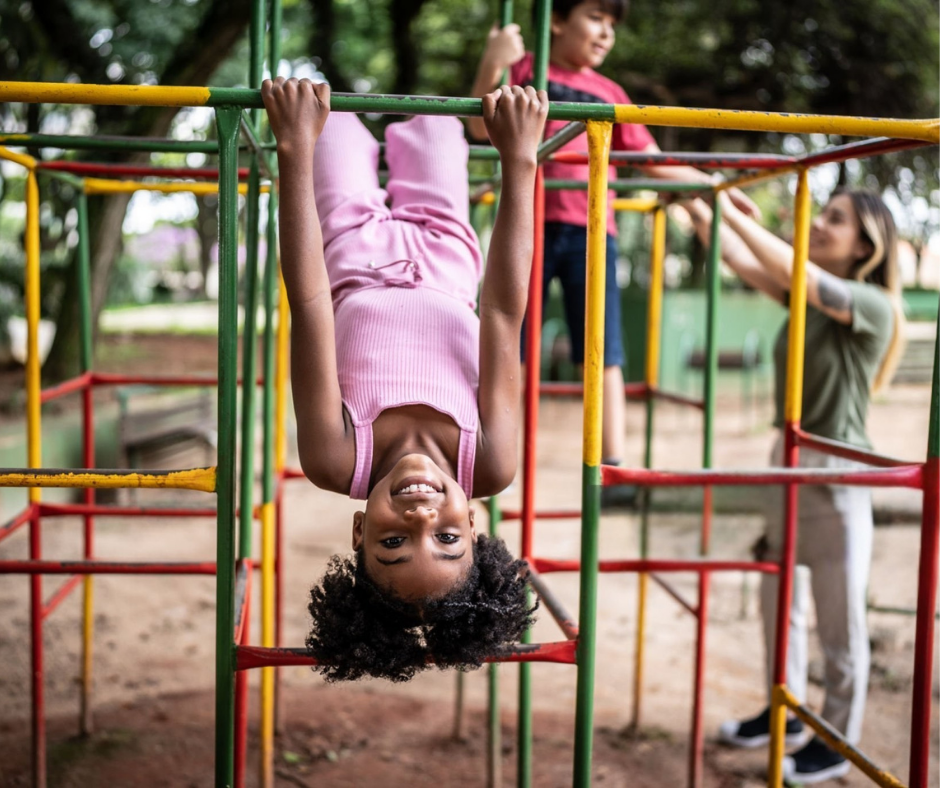 A young girl hanging upside down on playground equipment, smiling at the camera, with two other children playing in the background.