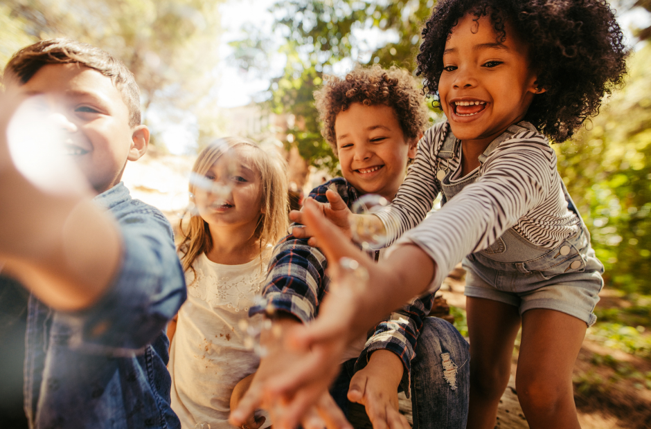 Four children playing outdoors in a sunny park, reaching for bubbles with excited expressions.