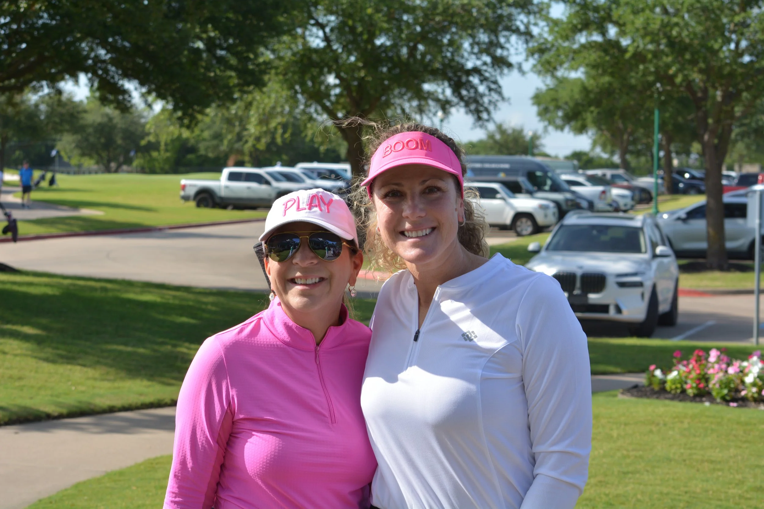 Two women smiling outdoors in sportswear, wearing pink and white hats, with a parking lot and trees in the background.