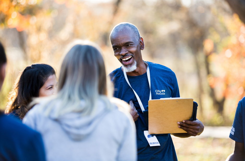 A smiling older man with a gray beard wearing a blue t-shirt holding a clipboard, talking to a group of young women outdoors with autumn trees in the background.