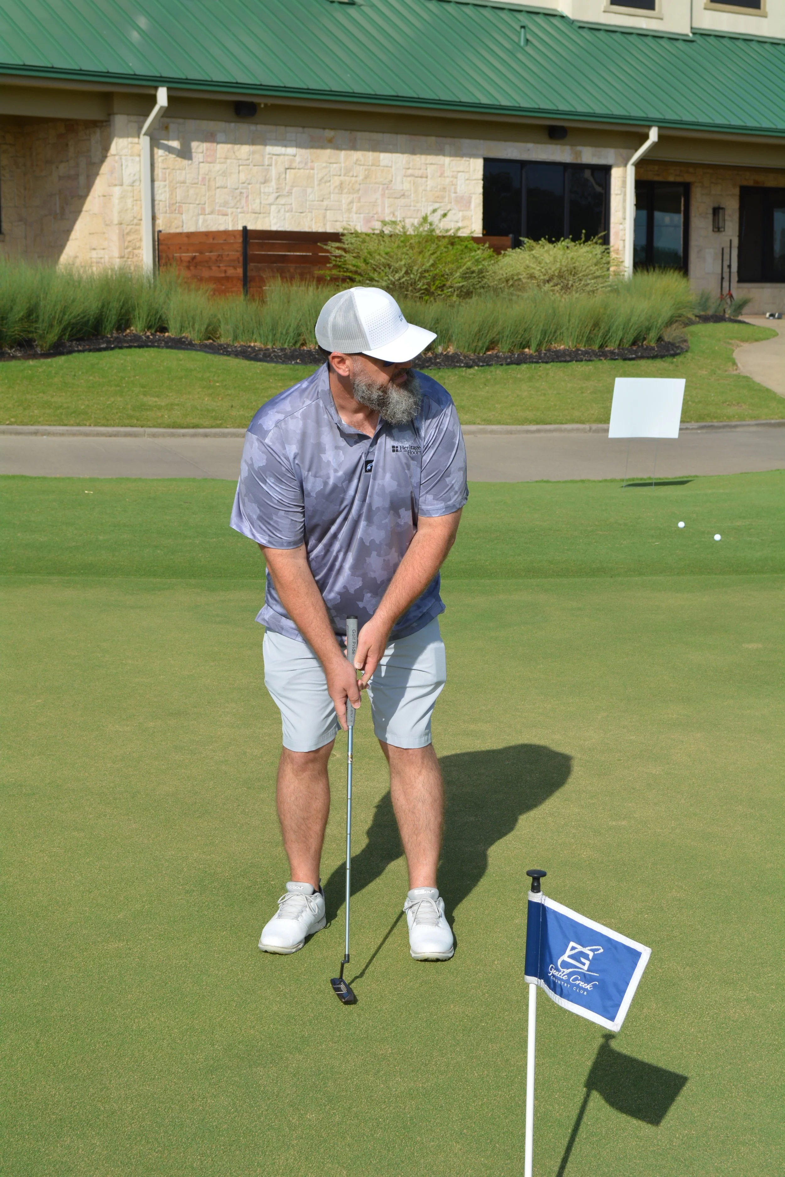 A man with a beard wearing a white cap, gray patterned polo shirt, and light shorts, stands on a golf green holding a golf club, preparing to putt on a sunny day.
