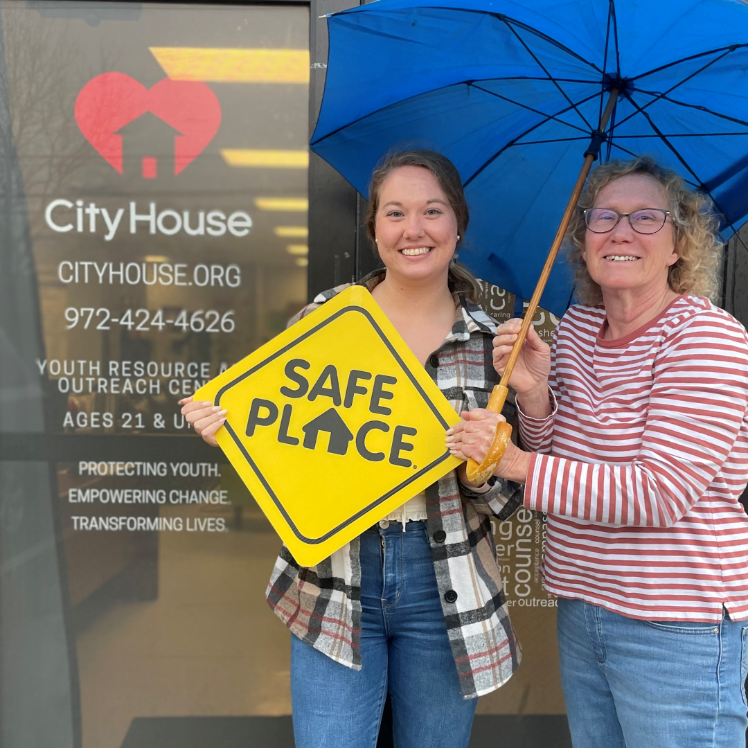 Two women standing outside a building with a sign that says 'City House' and holding a yellow sign that says 'Safe Place'. One woman is holding a blue umbrella.
