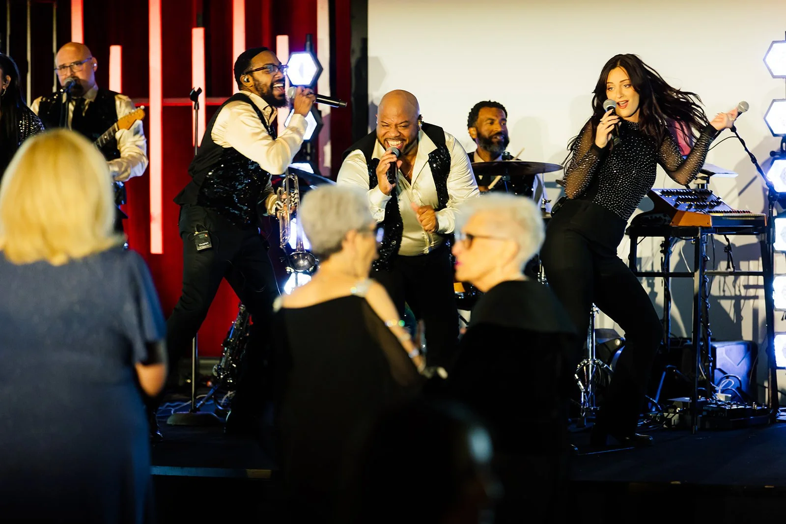 A live music performance with three singers singing and dancing on stage, surrounded by band members playing instruments, with an audience watching.