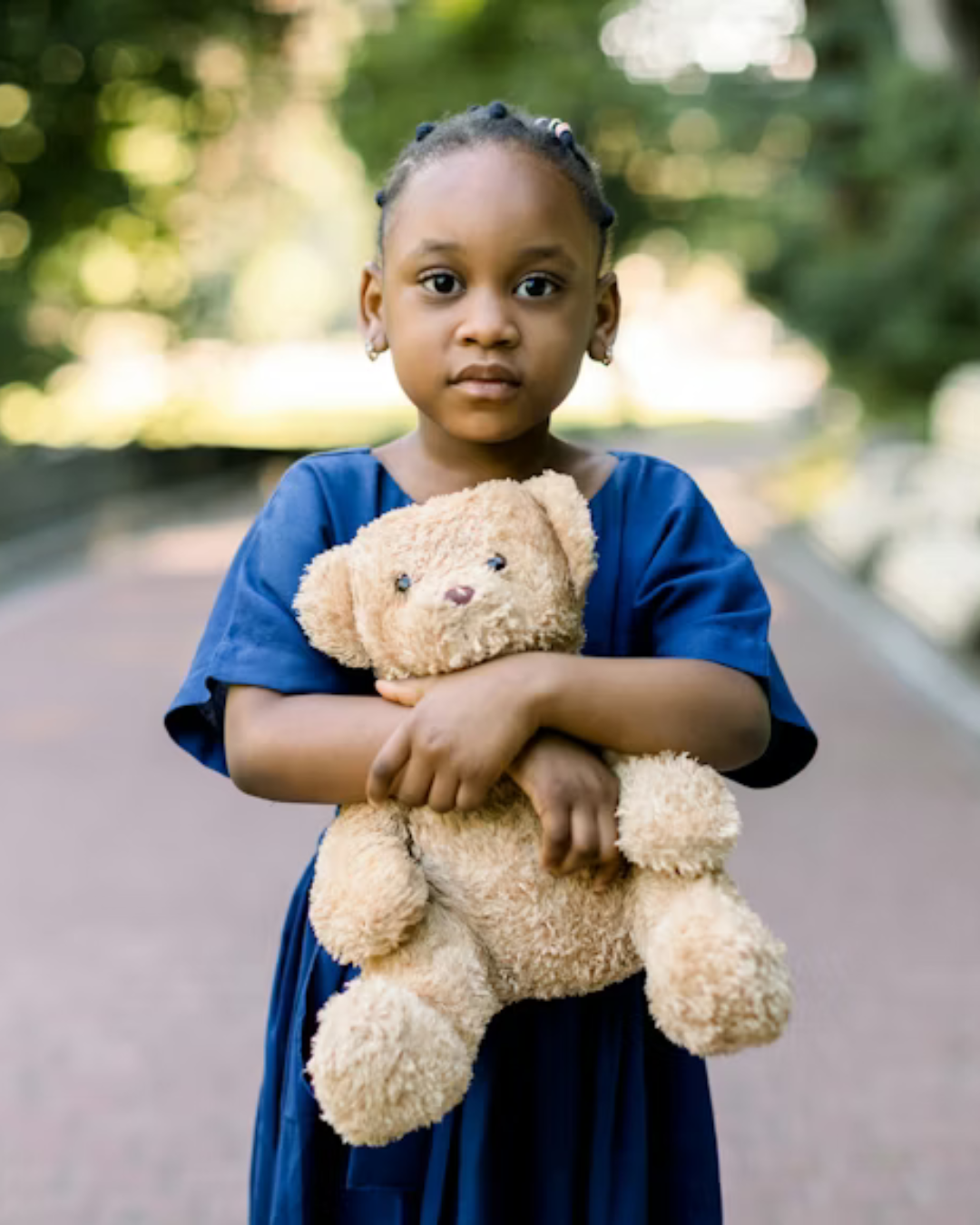 Young girl with braided hair and earrings wearing a blue dress, holding a plush teddy bear outdoors on a blurred green background.