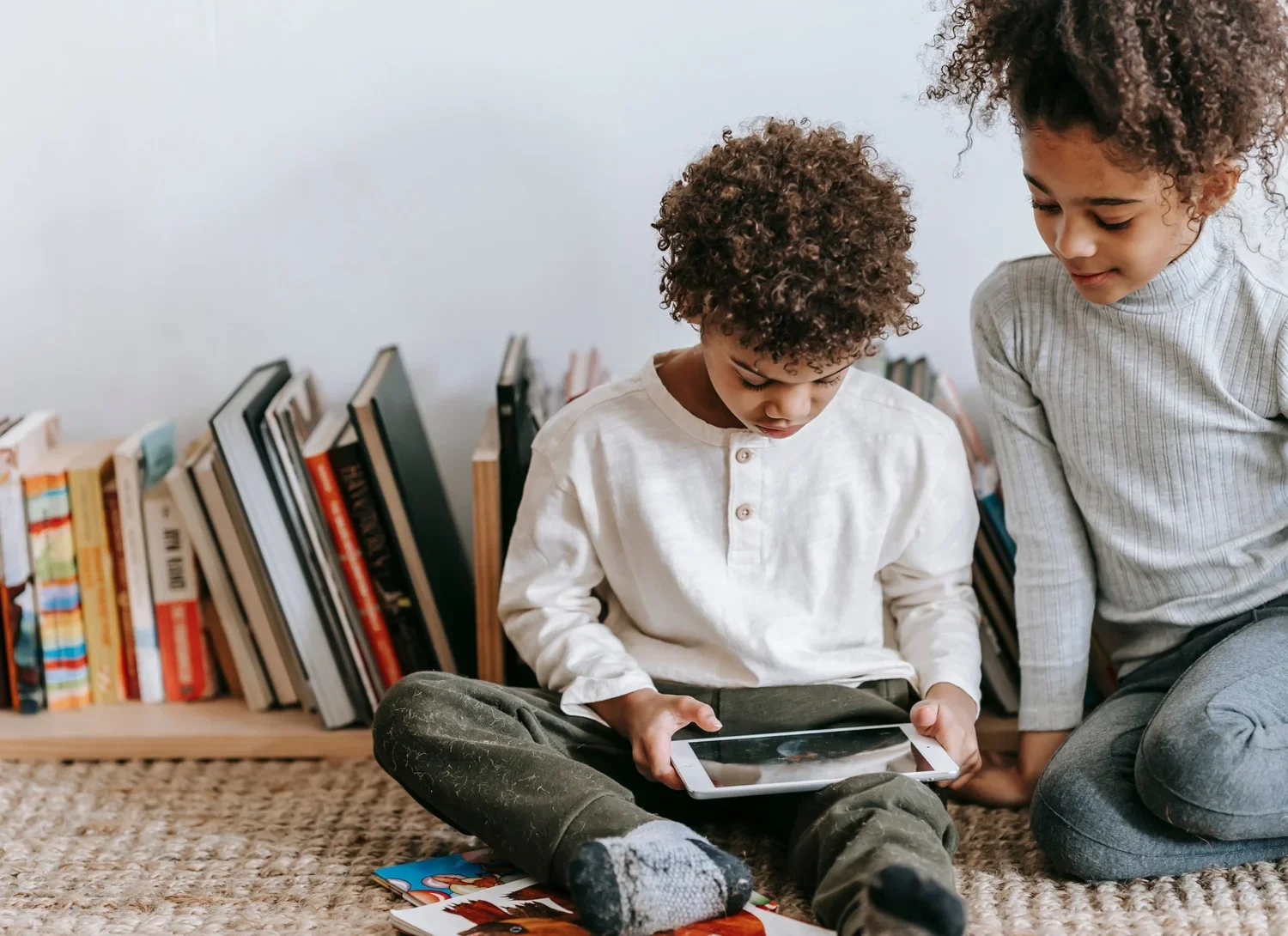 Two children, a boy and a girl, sitting on a carpet in front of a bookshelf, looking at a tablet together.