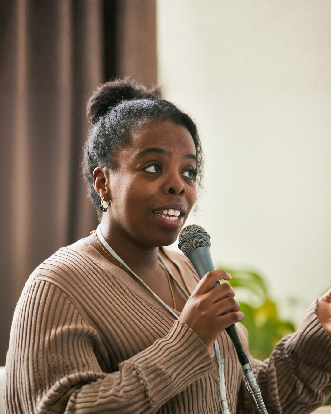 A young woman with her hair in a bun, wearing a beige ribbed sweater, speaking into a microphone indoors with a blurred background and green plants.