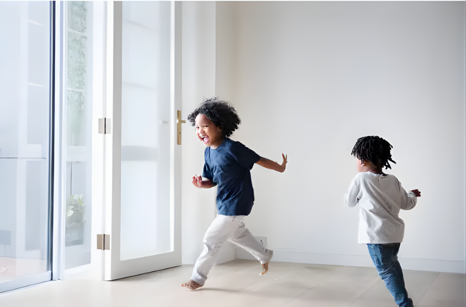 Two children playing and running inside a bright, minimalistic room near glass doors.
