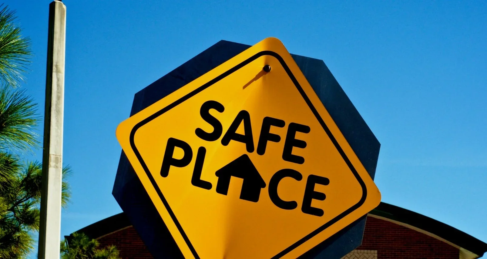 Yellow diamond-shaped sign with black border and text reading 'SAFE PLACE', mounted on a post against a blue sky, with a building and green trees in the background.