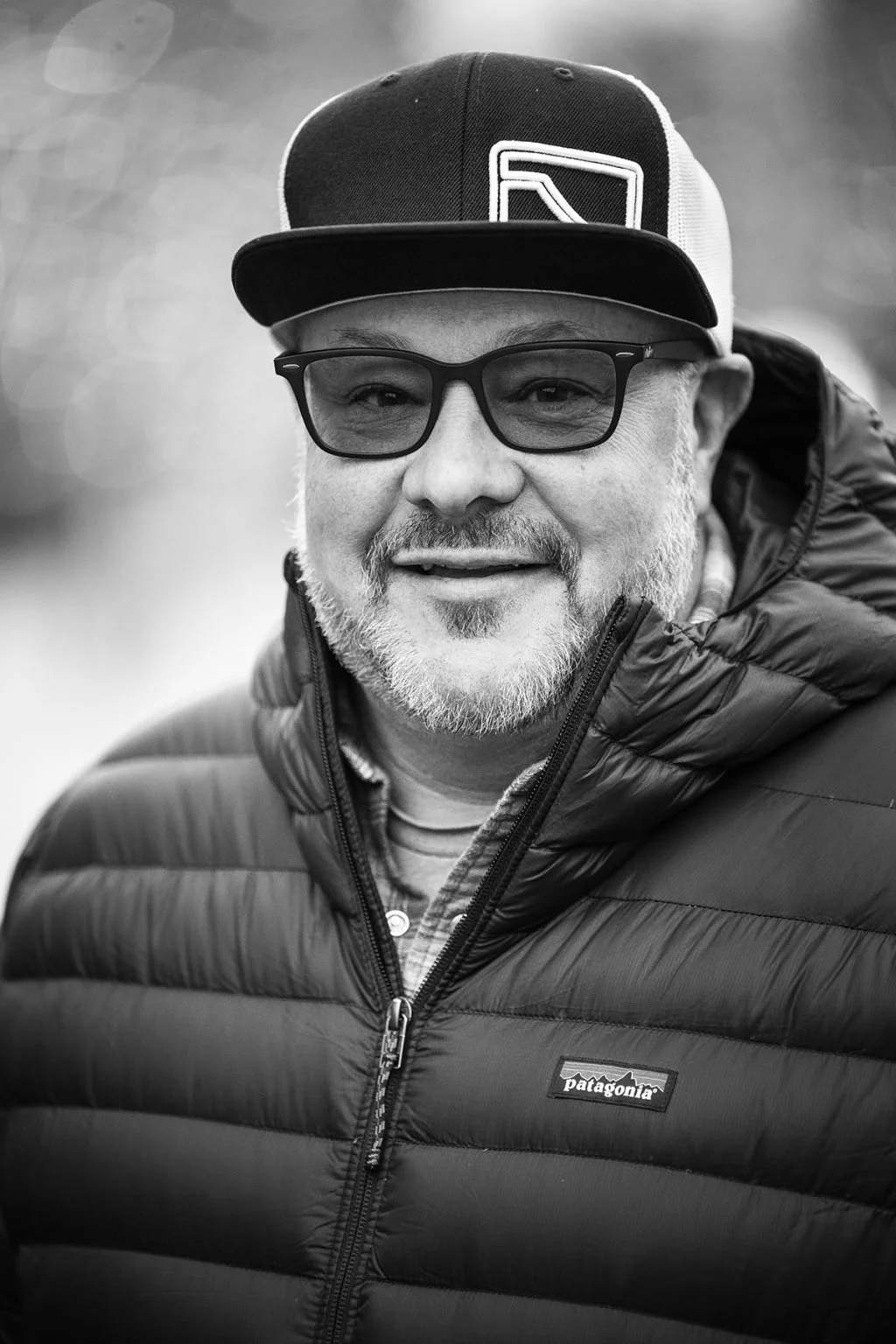 A middle-aged man with glasses, a beard, and a mustache smiling outdoors. He is wearing a baseball cap and a puffy Patagonia jacket.