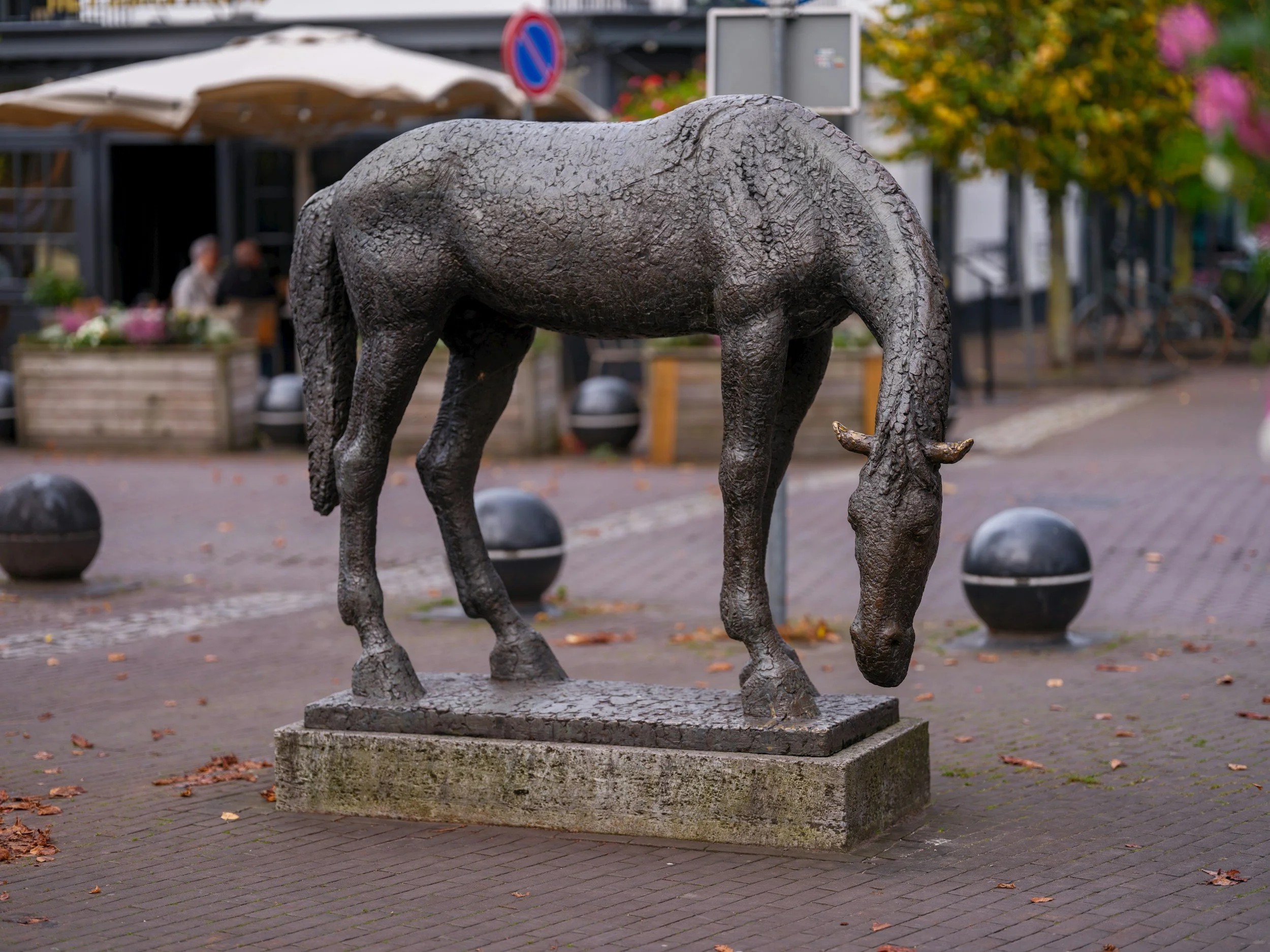 Bronze sculpture of a grazing horse on a paved sidewalk in an urban setting, with outdoor seating, people, and trees in the background.