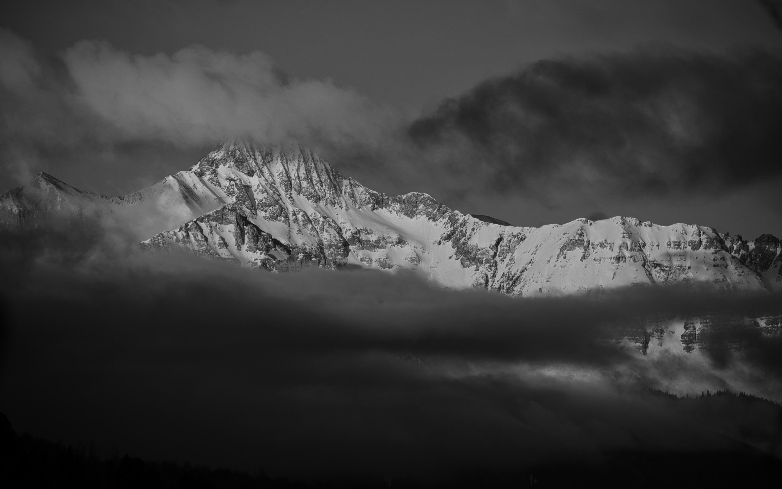 Black and white photograph of snow-covered mountain peaks shrouded in clouds and fog.