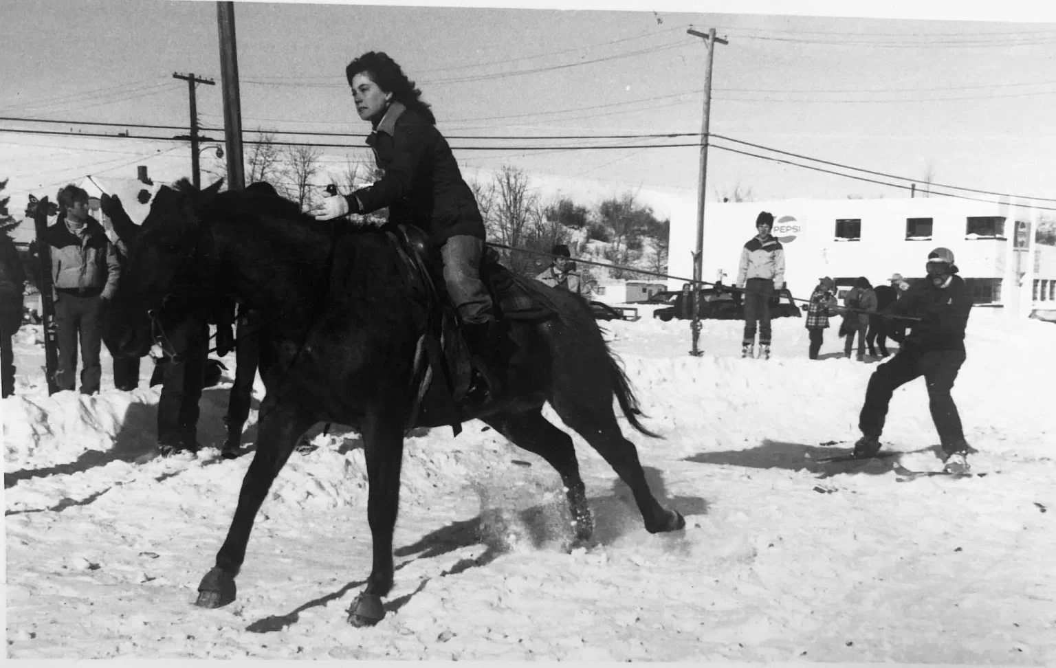 A black and white photo of a woman riding a horse in a snowy outdoor setting with several people in the background, some of whom are on skis and others standing.